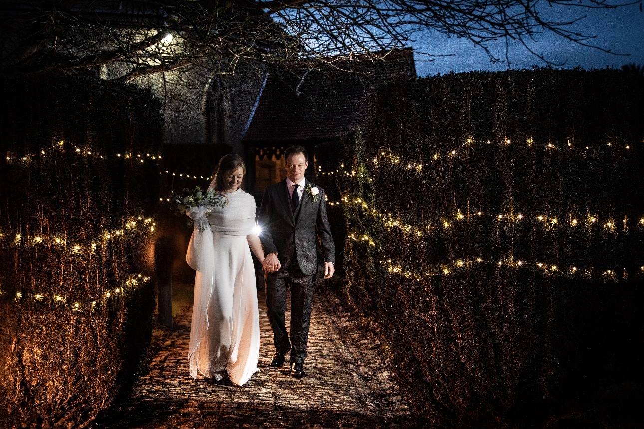 Wedded couple walk down path at dusk lit by fairylights
