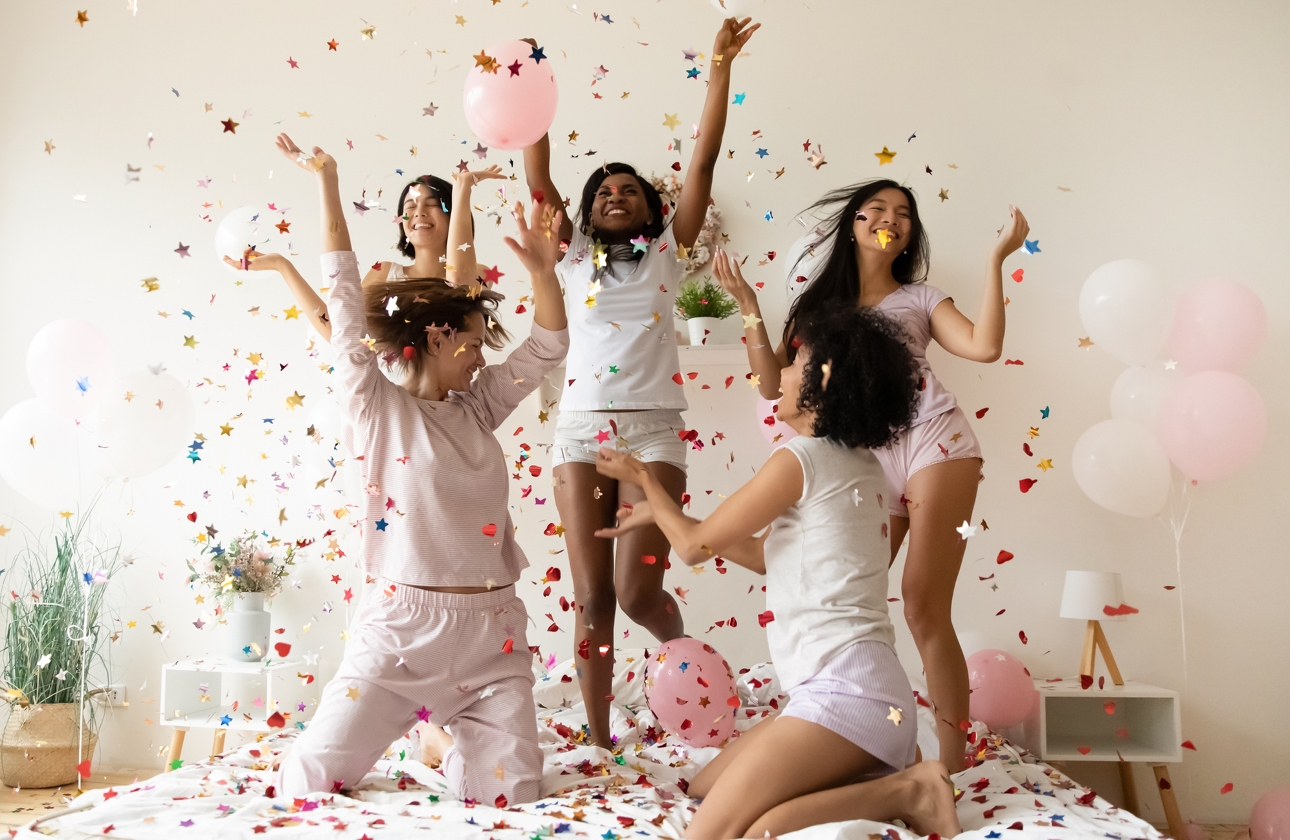 group of women on a bed jumping about with balloons and confetti